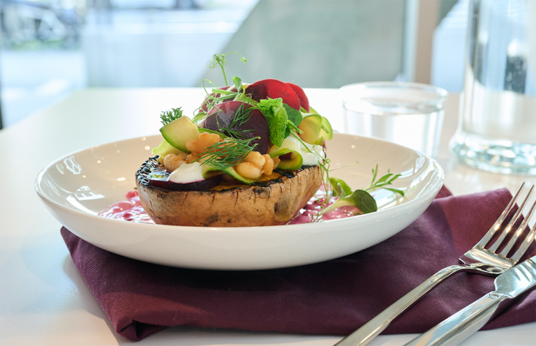 Ein Gourmet-Gericht mit einer großen gegrillten Pilzkappe, die mit Kichererbsen, Avocadoscheiben, Grünzeug und Sauce auf einem weißen Teller serviert wird, mit einer violetten Serviette, einem Glas Wasser, einer Gabel und einem Messer an der Seite.