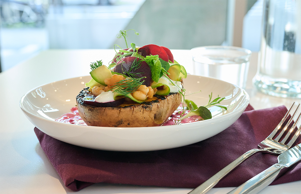 Ein Gourmet-Gericht mit einer großen gegrillten Pilzkappe, die mit Kichererbsen, Avocadoscheiben, Grünzeug und Sauce auf einem weißen Teller serviert wird, mit einer violetten Serviette, einem Glas Wasser, einer Gabel und einem Messer an der Seite.