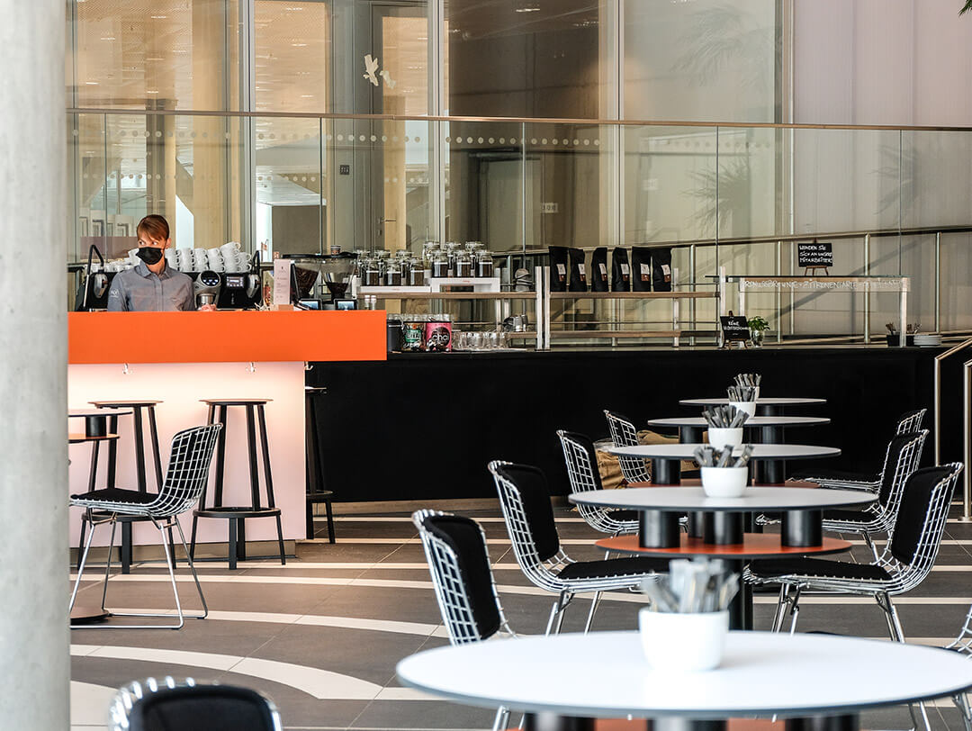 Ein modernes Café mit schwarzen und weißen Tischen und Stühlen, leer bis auf einen maskierten Mitarbeiter, der hinter dem orangefarbenen Tresen steht. Glaswände und natürliches Licht schaffen eine offene, luftige Atmosphäre.