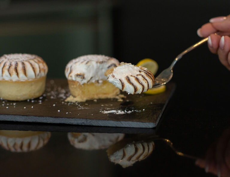 Nahaufnahme einer Hand, die einen Löffel mit einem Bissen Zitronen-Baiser-Torte hält, mit zwei Mini-Torten, die mit geröstetem Baiser und Puderzucker auf einer Schiefertafel im Hintergrund belegt sind.