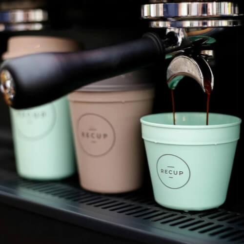 Nahaufnahme von Kaffee, der in einen hellgrünen wiederverwendbaren RECUP-Becher gebrüht wird, mit zwei weiteren wiederverwendbaren Bechern, einem grünen und einem beigen, im Hintergrund unter einer Espressomaschine.