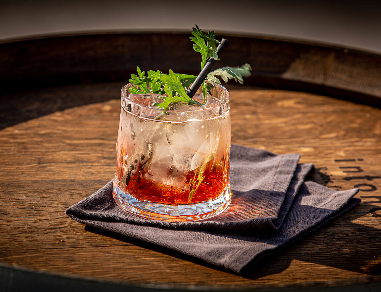 Ein Cocktail mit Eis und einer roten Flüssigkeit, garniert mit grünen Kräutern und serviert mit einem schwarzen Strohhalm, liegt auf einer dunklen Stoffserviette auf einer Holzfläche.