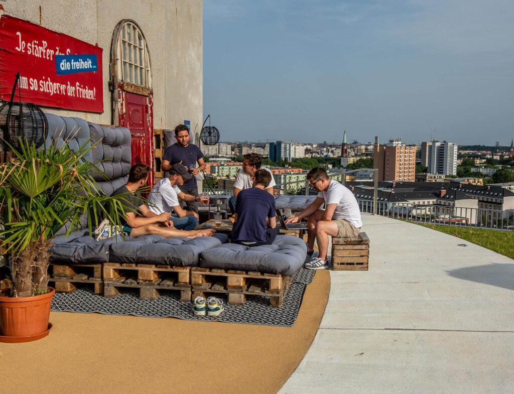 Eine Gruppe von sechs Personen sitzt auf gepolsterten Holzpaletten auf einer Dachterrasse, unterhält sich und entspannt sich. Im Hintergrund ist die Stadtlandschaft mit ihren Hochhäusern und dem klaren Himmel zu sehen. Eine Topfpflanze und ein Wandschild stehen in der Nähe.