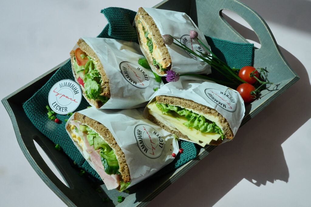 Ein Tablett mit vier eingewickelten Sandwiches, gefüllt mit Salat, Käse und Wurstwaren, daneben ein Zweig Kirschtomaten und Kräuter, auf einem dunklen Tuch, auf dem die Etiketten "MAMAMIA lecker" sichtbar sind.