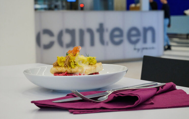 Auf einem Tisch steht ein geschirrter Gourmet-Teller mit Gemüse, daneben liegen Gabel und Messer auf einer lila Serviette. Im Hintergrund steht ein Schild mit der Aufschrift "Kantine" in einem modernen, hellen Ambiente.