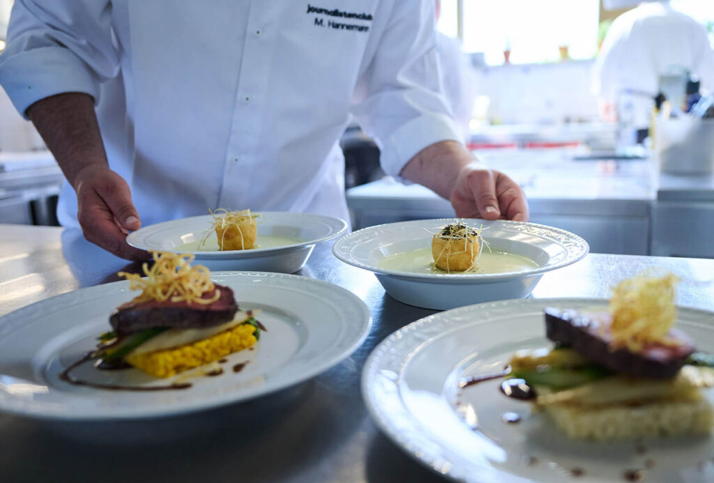 Ein Koch im weißen Kittel arrangiert elegante Teller mit Gourmetgerichten auf einem Tresen in einer hellen, modernen Küche. Die Gerichte sind mit kunstvoll angerichteten Fleisch- und Gemüsesorten und goldenen Garnierungen versehen.