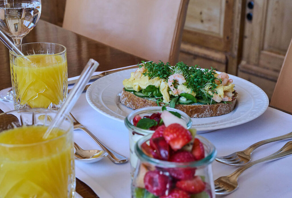 Ein Frühstückstisch mit Orangensaft, einem Teller mit einem belegten Brötchen mit Eiern, Krabben, Grünzeug und Kräutern und Gläsern mit frischem Obstsalat, dazu Besteck und Gläser.