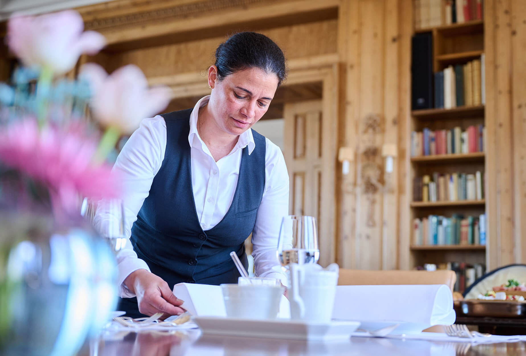 Eine Frau in weißem Hemd und dunkler Weste deckt einen Esstisch in einem gemütlichen, holzgetäfelten Raum mit Bücherregalen und Blumen im Vordergrund.