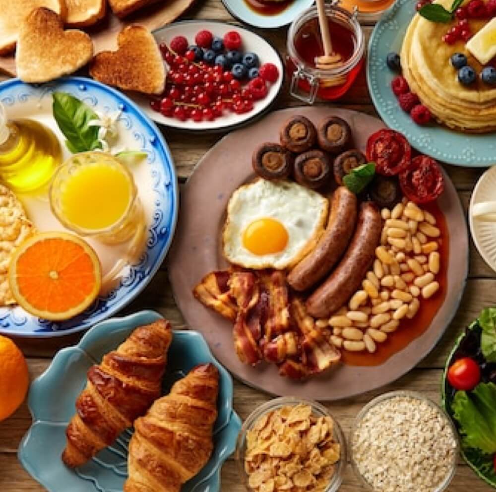 Eine Vielzahl von Frühstücksspeisen auf einem Tisch, darunter Pfannkuchen mit Beeren, Toast, Saft, Croissants, Müsli, ein komplettes englisches Frühstück mit Eiern, Speck, Würstchen, Bohnen, Pilzen und Tomaten sowie Obst und Honig.