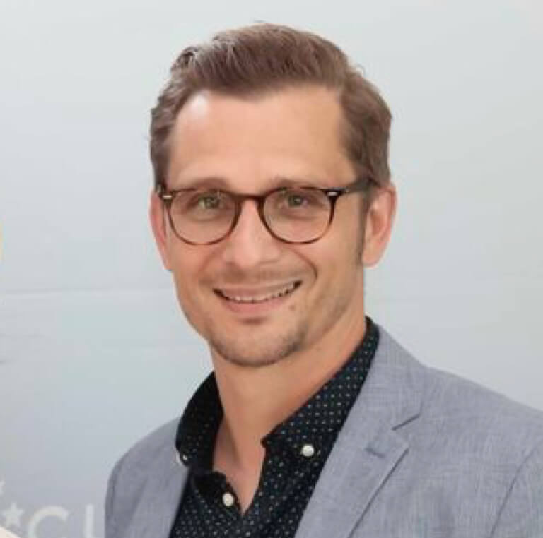 Ein Mann mit kurzem, hellbraunem Haar und Brille, der einen hellgrauen Blazer über einem dunklen, gemusterten Hemd trägt, lächelt in die Kamera vor einem hellen Hintergrund.