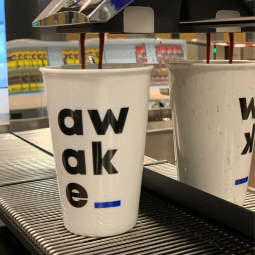 Eine weiße Tasse mit dem Wort "awake" in fetten schwarzen Buchstaben wird mit Kaffee aus einer Maschine gefüllt. Die Reflexion der Tasse ist auf der Metalloberfläche daneben sichtbar.