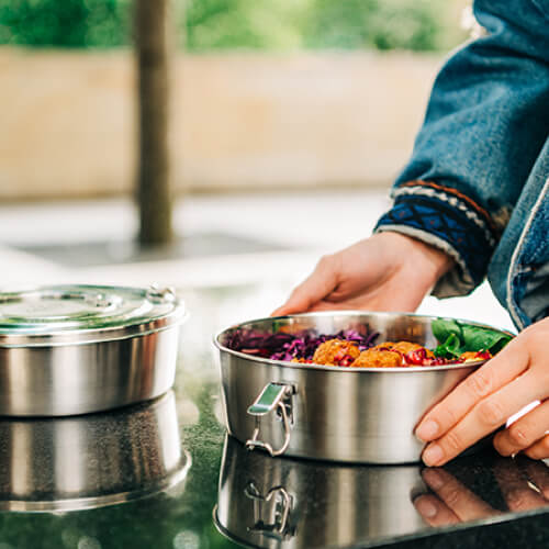 Eine Person in Jeansjacke hält eine runde Lunchbox aus Edelstahl, die mit bunten Lebensmitteln gefüllt ist, neben einem anderen geschlossenen Behälter auf einer glänzenden Außenfläche.