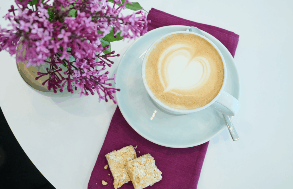Eine Tasse Cappuccino mit herzförmigem Schaum sitzt auf einer weißen Untertasse mit Löffel, daneben zwei kleine Gebäckstücke und eine Vase mit lila Blumen auf einer lila Serviette.