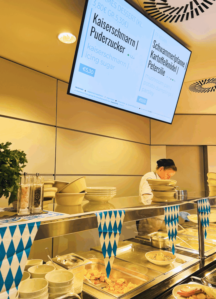 Eine mit blau-weißen Wimpeln, Geschirr und Kräutern geschmückte Cafeteria-Theke erinnert an das Oktoberfest, während ein Koch - vielleicht PAPA genannt - das Essen serviert. Auf einer großen Speisekarte stehen Kaiserschmarrn mit Puderzucker und Kartoffelpuffer mit Petersilie.