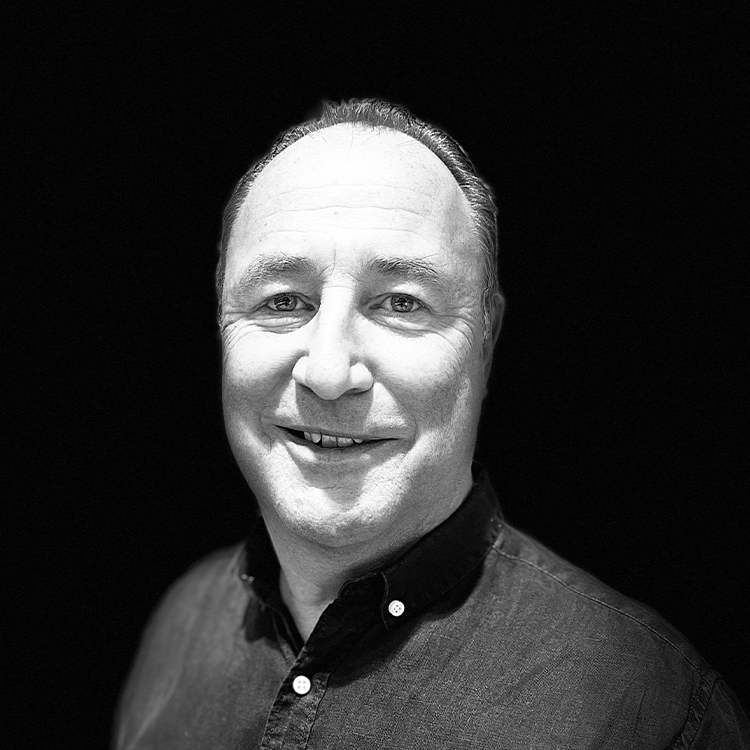 Schwarz-Weiß-Porträt von Lars Tretzack, einem Mann mittleren Alters mit kurzen Haaren, der ein zugeknöpftes Hemd trägt, lächelt und leicht nach links blickt, vor einem schlichten schwarzen Hintergrund.