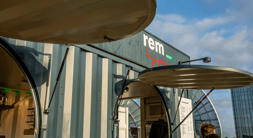 Nahaufnahme eines modernen Imbisswagens aus einem Schiffscontainer mit der Aufschrift REM, mit offenen Servicefenstern und einer sichtbaren Speisekarte. Die sonnige Szene mit blauem Himmel deutet auf den Saisonabschluss hin, der die Feiertagspause für dieses einzigartige Lokal markiert.