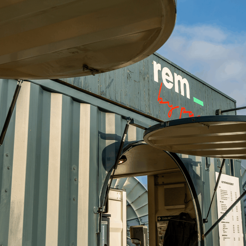 Nahaufnahme eines modernen Imbisswagens aus einem Schiffscontainer mit der Aufschrift REM, mit offenen Servicefenstern und einer sichtbaren Speisekarte. Die sonnige Szene mit blauem Himmel deutet auf den Saisonabschluss hin, der die Feiertagspause für dieses einzigartige Lokal markiert.