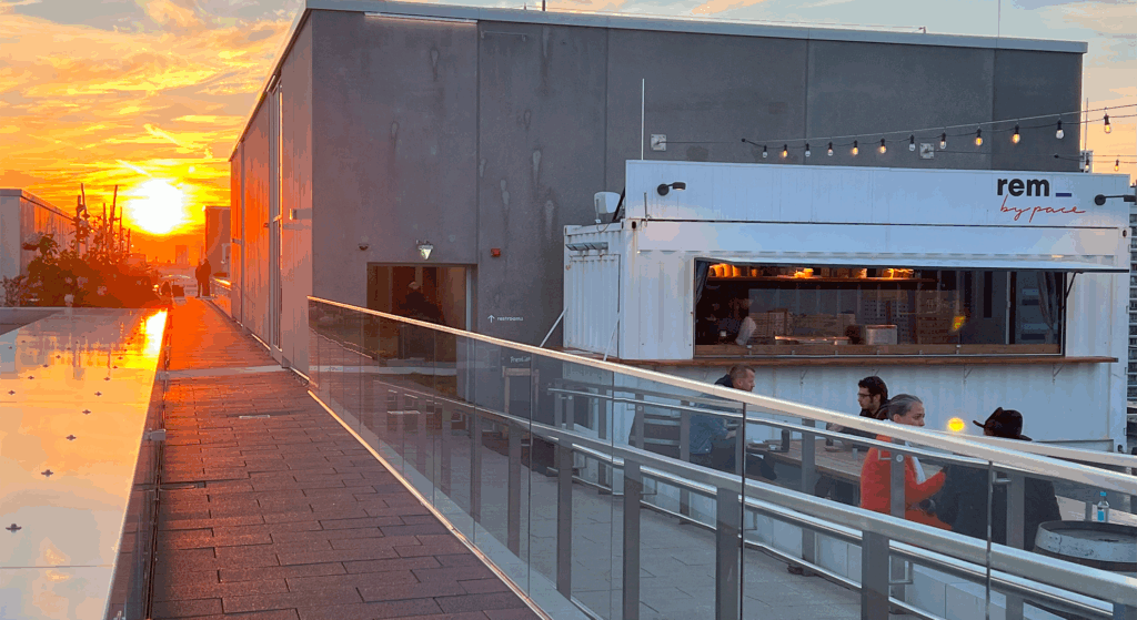 Eine Rooftop-Bar bei Sonnenuntergang mit einem modernen grauen Gebäude, Glasgeländern und Menschen neben einem weißen Container mit der Aufschrift "REM by pace". Das warme Sonnenlicht spiegelt sich auf den Oberflächen und schafft eine lebendige, gemütliche Atmosphäre - perfekt für einen Saisonabschluss oder eine Feiertagspause.