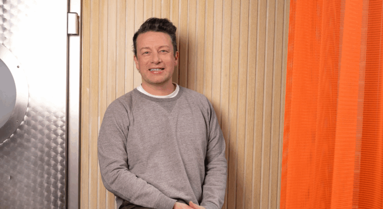 Ein Mann mit kurzen dunklen Haaren, der ein graues Sweatshirt und ein weißes Unterhemd trägt, steht vor einer hellen Holzwand in der Nähe eines orangefarbenen Vorhangs und einer CANTEEN-Metalltür und lächelt in die Kamera - er verkörpert Jamie Olivers Leidenschaft für gesundes Essen.