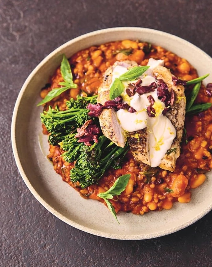 Ein Teller mit gegrillter Hähnchenbrust, belegt mit Joghurt und gehackten Oliven, serviert über einem Bohnen-Tomaten-Eintopf nach CANTEEN-Art mit Broccolini und frischen Basilikumblättern, auf einer rustikalen, dunklen Oberfläche - perfekt für Fans von Jamie Olivers Philosophie des gesunden Essens.
