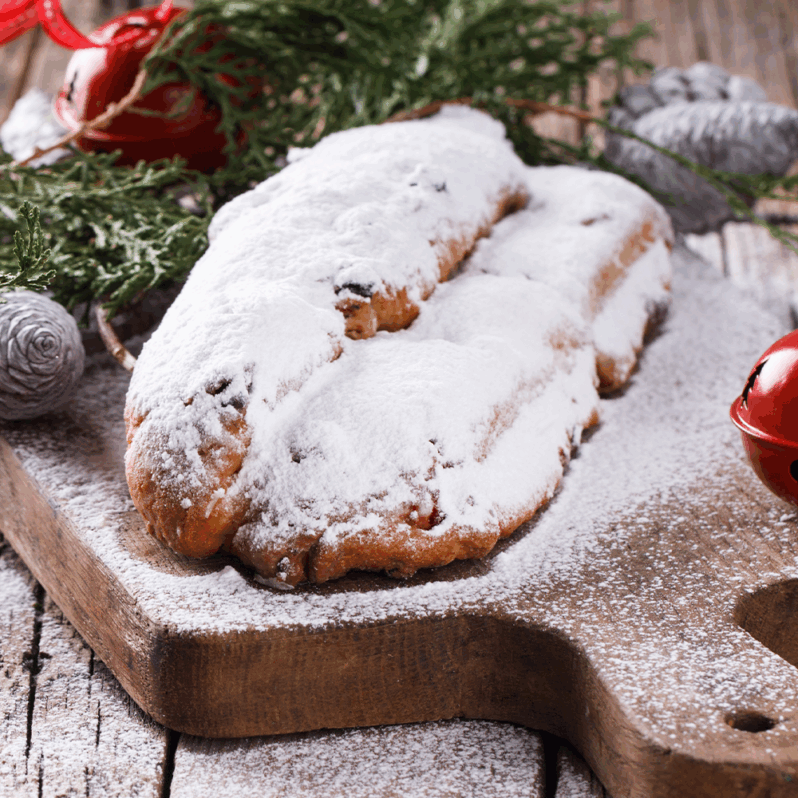 Zwei Stücke süßer, saftiger Stollen, bestäubt mit Puderzucker, liegen auf einem hölzernen Schneidebrett, umgeben von immergrünen Zweigen und roten Jingle Bells als festliche Weihnachtsdekoration im Hintergrund.