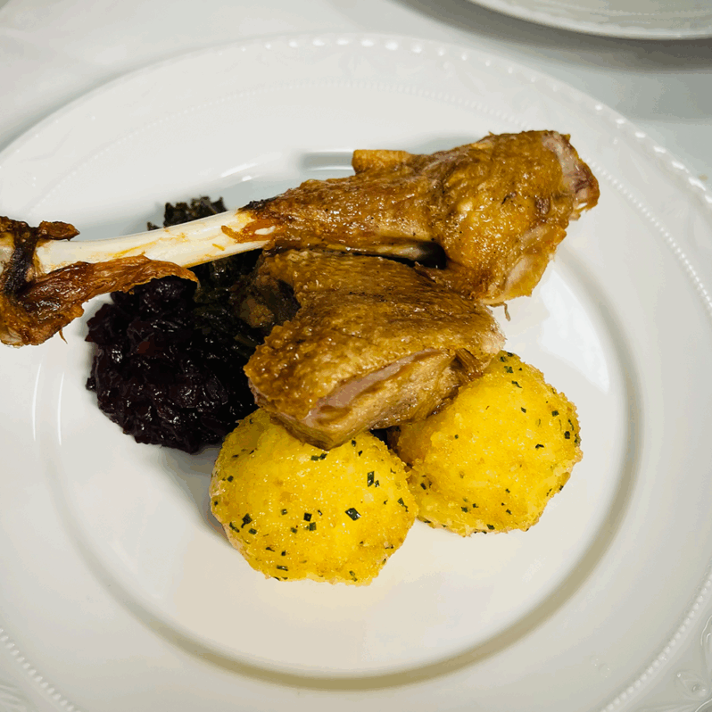 Ein Tellergericht für ein festliches Dinner: gebratene Entenkeule, zwei runde gelbe Knödel mit Kräutern, Rotkohl und Grünzeug, alles elegant angerichtet auf einem weißen Dekoteller - perfekt für einen besonderen Abend im JOURNALISTENCLUB.