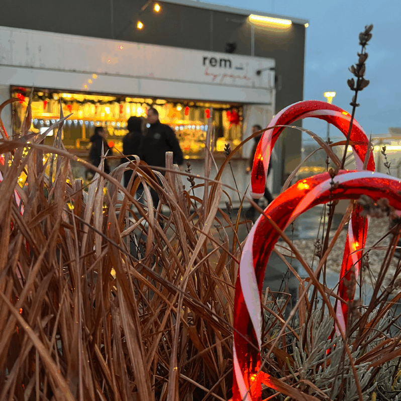 Zwei beleuchtete Zuckerstangendekorationen stehen zwischen getrocknetem Gras im Vordergrund, während im Hintergrund ein festlicher Weihnachtsmarktstand und verschwommene Menschen zu sehen sind. Warme Lichter schaffen eine gemütliche Atmosphäre voller Wintervergnügen.