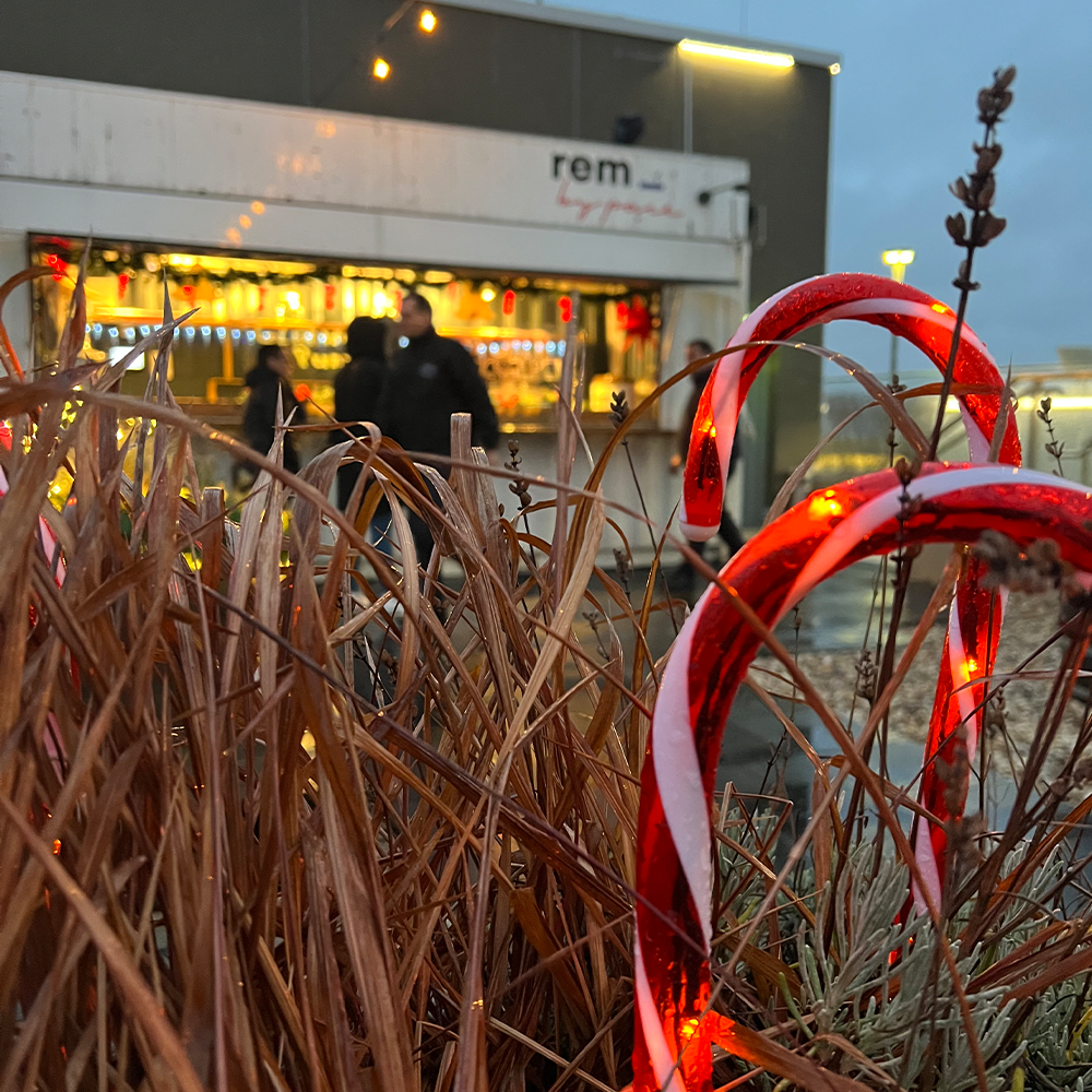 Zwei beleuchtete Zuckerstangendekorationen stehen zwischen getrocknetem Gras im Vordergrund, während im Hintergrund ein festlicher Weihnachtsmarktstand und verschwommene Menschen zu sehen sind. Warme Lichter schaffen eine gemütliche Atmosphäre voller Wintervergnügen.