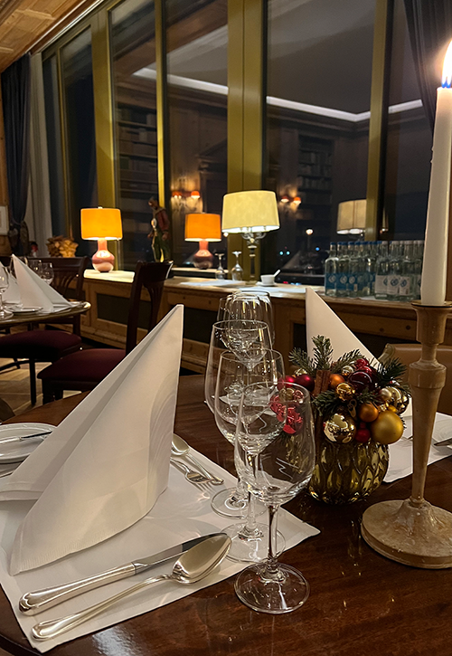 A cozy restaurant table is set for a festive dinner with folded white napkins, glasses, and cutlery. A burning candle and small festive decorations adorn the table in the JOURNALISTENCLUB. Warm lamps shine near the large windows at night.