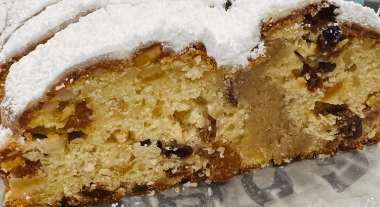 Eine Nahaufnahme eines aufgeschnittenen und mit Puderzucker bestäubten Stollens, der eine süße, saftige Textur aufweist, die mit Nüssen, Trockenfrüchten und einer Marzipanfüllung in der Mitte gefüllt ist.