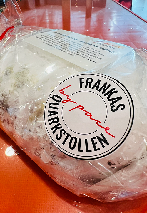 Ein Laib süßer, saftiger Frankas-Quarkstollen, eingewickelt in klarem Plastik mit einem weiß-schwarzen Etikett, liegt auf einer orangefarbenen Fläche. Auf dem Etikett steht "Frankas by pace Quarkstollen".
