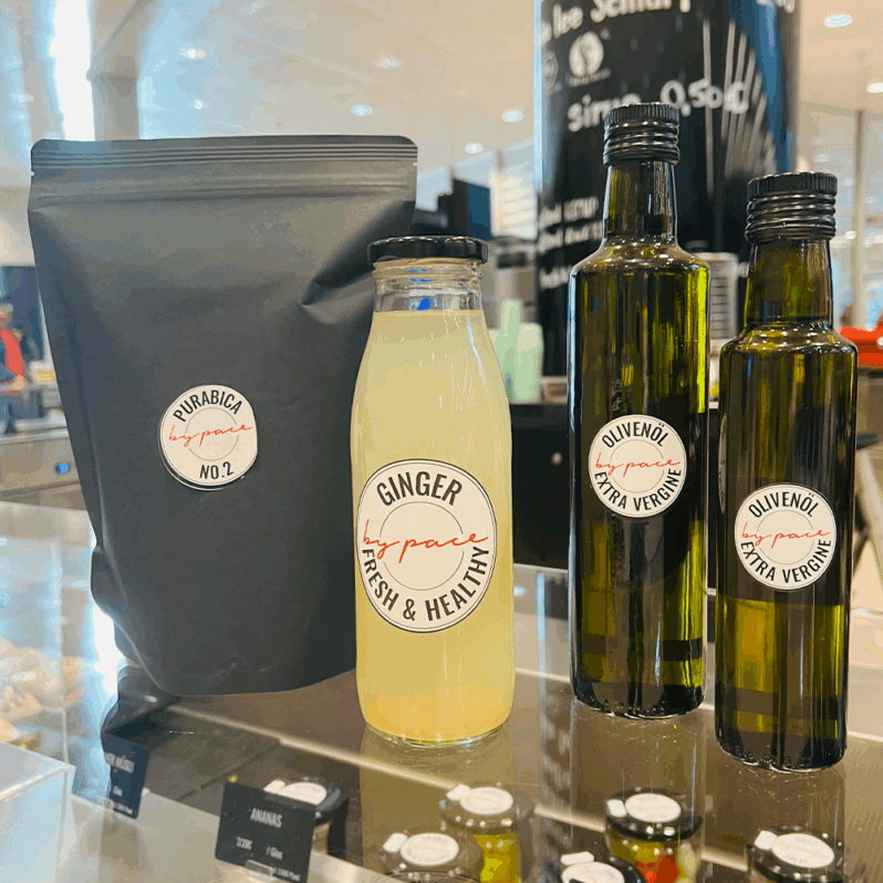 Drei Produkte auf einer Theke: Furrabica No. 2-Kaffee, eine Glasflasche Ingwerdrink - perfekt für die kalte Jahreszeit - und zwei dunkle Flaschen natives Olivenöl extra, alle mit weißen, handgeschriebenen Etiketten, die in jede stilvolle Küche passen.