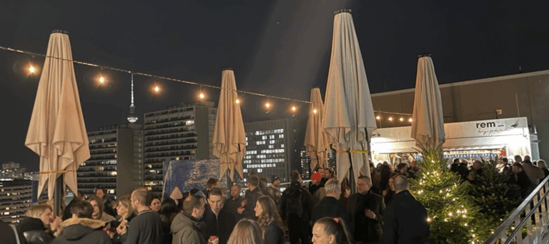 Eine große Gruppe von Menschen versammelt sich nachts auf einem Dach, umgeben von geschlossenen Schirmen, Lichterketten und einem Weihnachtsbaum, und genießt das an einen Weihnachtsmarkt erinnernde Vergnügen, mit städtischen Gebäuden und einem beleuchteten Fernsehturm im Hintergrund.