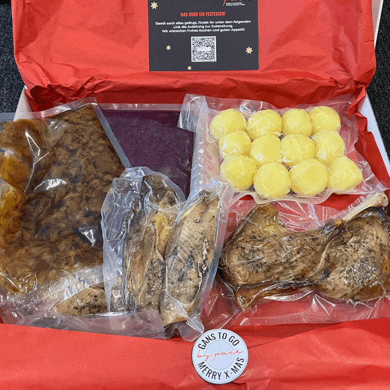 Ein himmlisches Mahlzeitenset in einer roten Schachtel mit vakuumverpackter Gans, herzhafter Soße und mehreren Runden gelber Knödel. Die Schachtel ist mit einem festlichen "Gans to go, Merry X-Mas"-Aufkleber versehen - ein perfekter Festtagsgenuss.