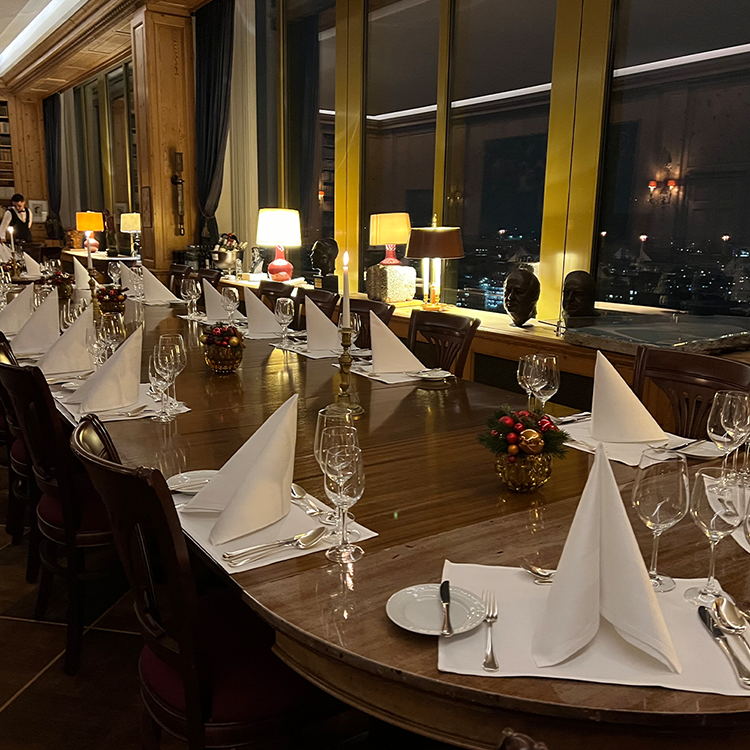 An elegantly set dining table for a goose dinner with folded white napkins, wine glasses, and festive centerpieces in the warmly lit JOURNALISTENCLUB, overlooking the nighttime cityscape through large windows.