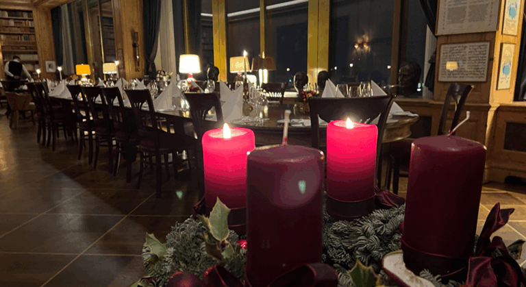 Eine Nahaufnahme von vier roten Kerzen und festlichem Grün, mit einem langen, für ein festliches Dinner gedeckten Esstisch in einem warm beleuchteten, eleganten, von Bücherregalen gesäumten Raum im JOURNALISTENCLUB.