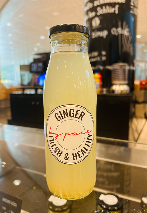 Eine Klarglasflasche mit schwarzem Deckel, gefüllt mit hellgelbem Ingwersaft. Auf dem Etikett steht Ginger von PACE Fresh & Healthy. Perfekt um fit zu bleiben, besonders in der kalten Jahreszeit. Die Flasche ist in einem Innenraum mit unscharfem Hintergrund ausgestellt.