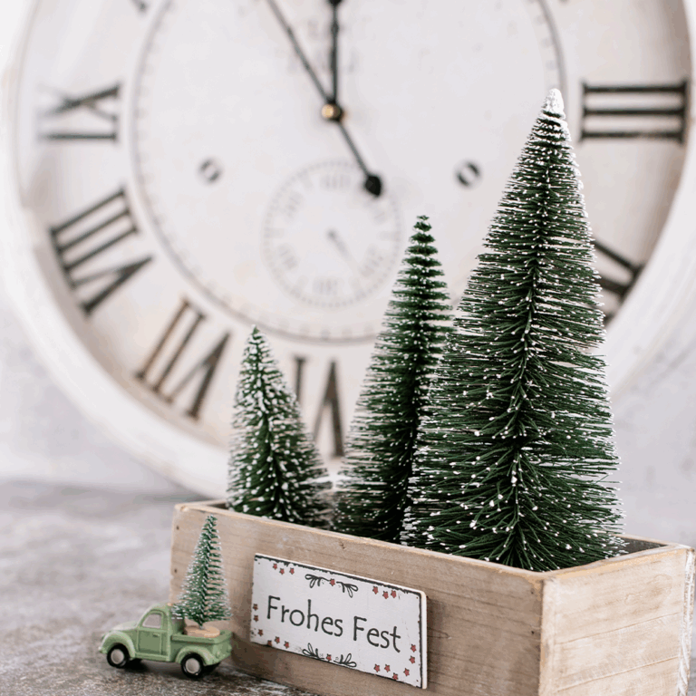 Eine Holzkiste mit kleinen künstlichen Weihnachtsbäumen und einem Schild mit der Aufschrift Frohes Fest steht vor einer großen Uhr und weist auf die Öffnungszeiten und die festliche Stimmung während der Feiertage hin. Neben der Kiste steht ein grüner Spielzeuglastwagen mit einem kleinen Baum.