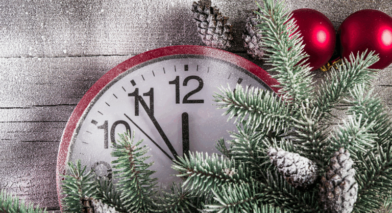 Eine Uhr, die fünf Minuten vor Mitternacht anzeigt, ist umgeben von verschneiten Tannenzweigen, Tannenzapfen und rotem Weihnachtsschmuck vor einem hölzernen Hintergrund, der eine festliche Jahreswechsel-Atmosphäre für die Feiertage hervorruft.