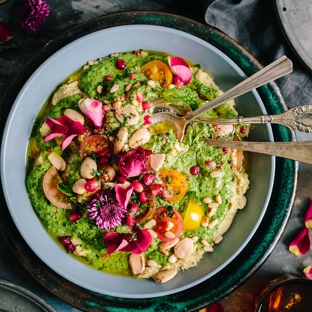 Eine Schale Hummus mit grüner Soße, Kirschtomaten, Nüssen, Samen, Granatapfelkernen und essbaren Blüten - dieses vegane Lieblingsgericht ist farbenfroh und kunstvoll garniert, perfekt für alle, die während des Veganuary 2026 veganes Kochen genießen.