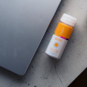 A silver laptop sits on a gray surface next to a small orange and white BOOST bottle, which claims to support daily energy levels and provide an energy boost for the whole day.
