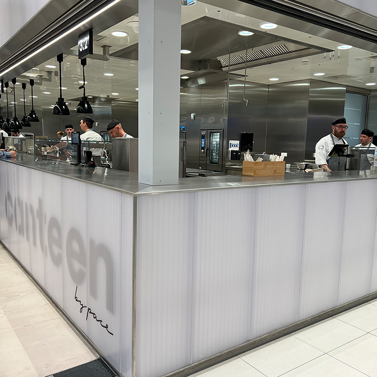 Eine moderne Cafeteria, in der Köche und Mitarbeiter hinter einem Tresen das Essen zubereiten. Die Kantine von ran bietet Einblicke in die Küche, mit einem sauberen, hellen Bereich mit Edelstahlgeräten und einem modernen Design.