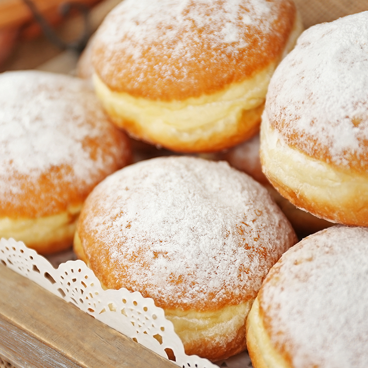 Eine Nahaufnahme mehrerer runder, mit Puderzucker überzogener Gelee-Donuts - bekannt als Pfannkuchen - in einem Holztablett, das mit einem dekorativen Papierdeckchen ausgelegt ist.