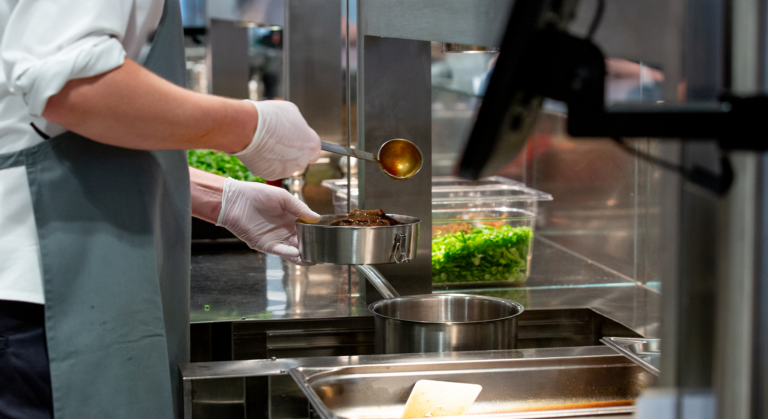 Hinter den Kulissen einer belebten Kantine schöpft eine Person mit Handschuhen und Schürze Soße auf das Essen in einer Metallschüssel, während in der Nähe gehacktes Grünzeug und andere Utensilien zu sehen sind.