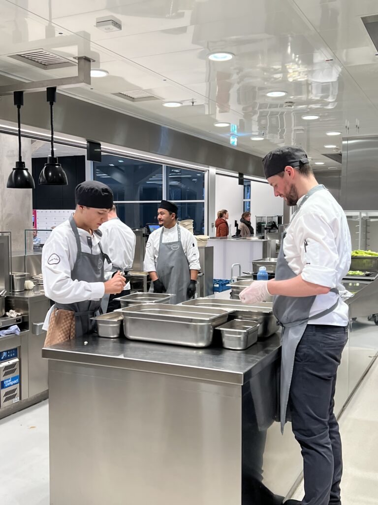 Vier Köche in weißen Uniformen und grauen Schürzen arbeiten in einer modernen Großküche zusammen und bereiten an einer Edelstahltheke das Essen zu. Dieser hell erleuchtete Raum bietet mit seinen großen Fenstern, die den Blick auf BLICK HINTER DIE KULISSEN freigeben, echte KÜCHENEINBLICKE.