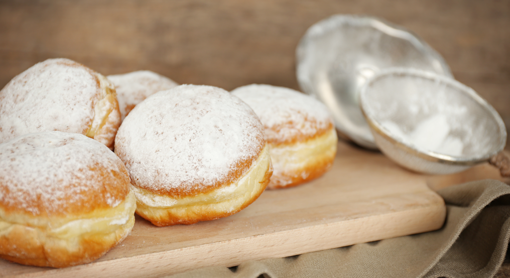 Fünf Puderzucker-Donuts liegen auf einem Holzbrett neben einem Sieb und einem beigen Tuch, das an klassische Pfannkuchen erinnert, mit einem rustikalen Holzhintergrund.