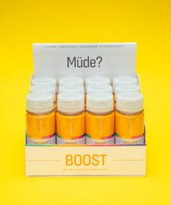 A box containing twelve orange BOOST drink bottles stands in front of a yellow background. A sign inside the box reads Müde? (German for tired?). The box bears the inscription BOOST BY NEUROSCIENTISTS – Your source for daily brain food and energy boosts.