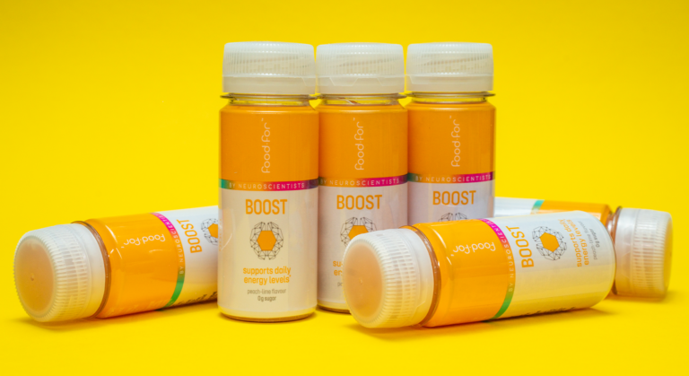 Sechs kleine Flaschen mit der Aufschrift BOOST von Food for Good, mit orangefarbenen und weißen Etiketten, sind auf einem leuchtend gelben Hintergrund angeordnet. Dieses Brainfood-Produkt soll das tägliche Energieniveau für ganztägige Energiekicks unterstützen.