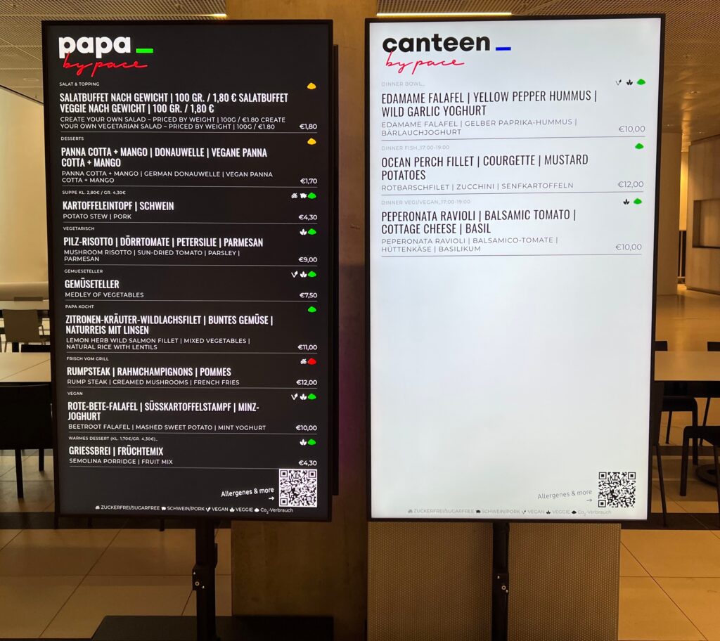 Zwei digitale Menütafeln zeigen das Essensangebot in einer Cafeteria an. Die linke Tafel, "papa by pace", zeigt Gerichte mit Preisen an. Die rechte, "canteen by pace", bietet leichtere Gerichte und Salate für nachhaltige Ernährung. Beide sind unten mit QR-Codes versehen.
