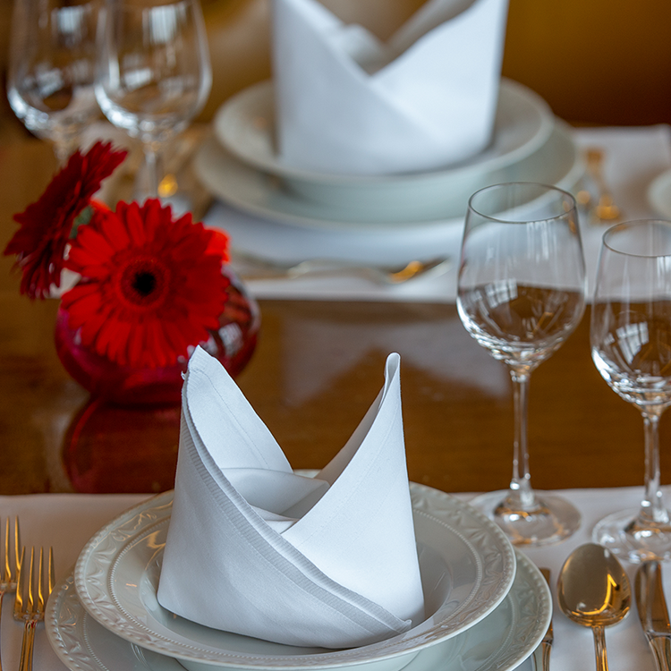 Eine Nahaufnahme eines elegant gedeckten Tisches beim Spargel-Dinner des JOURNALISTENCLUBs, mit weißen gefalteten Servietten auf Tellern, Weingläsern, Silberbesteck und einer kleinen roten Vase mit zwei roten Blumen - in einer formellen, einladenden Atmosphäre mit Blick auf die Skyline.